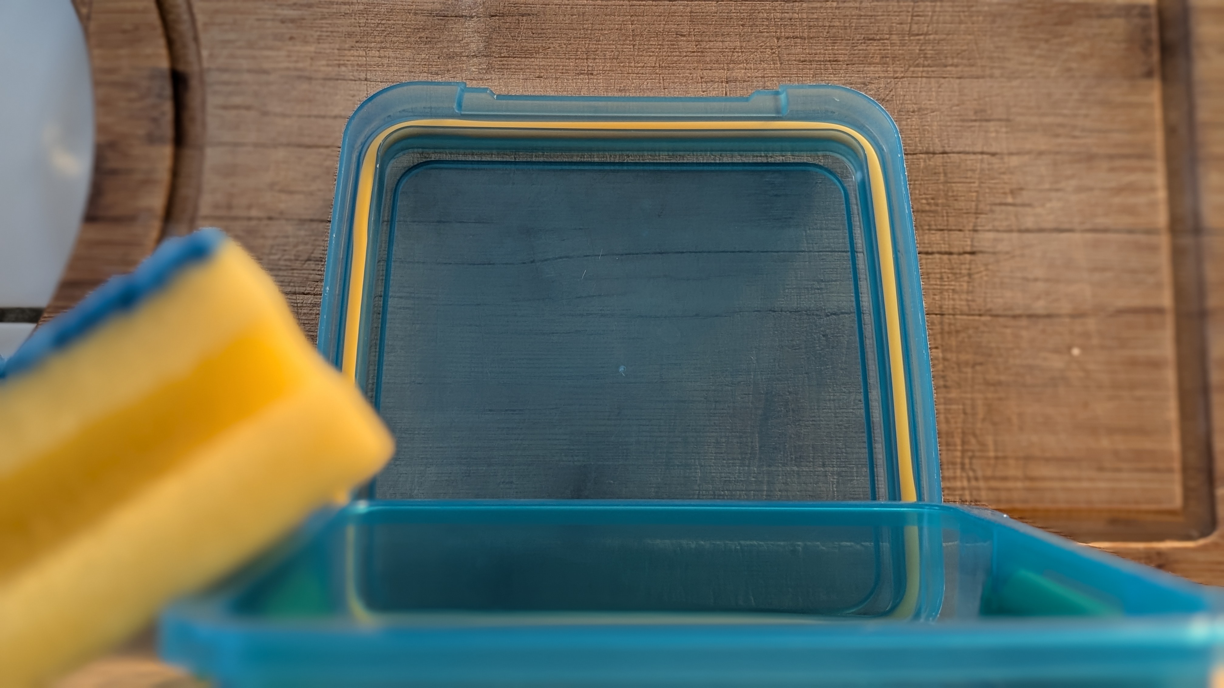 Das Bild zeigt eine geöffnete blaue Plastikbox mit gelben Akzenten, die auf einer Holzoberfläche steht. Links im Bild ist ein gelber Schwamm zu sehen, der teilweise unscharf ist.